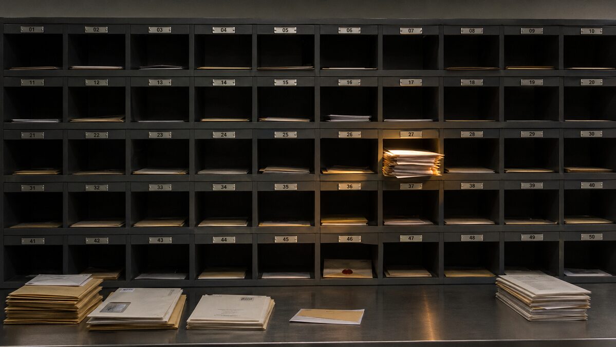 A long horizontal wall of identical numbered metal PO-box doors in a private mail-forwarding office, most sealed flush, with one box near the centre slightly ajar and lit from inside in warm amber, correspondence protruding.