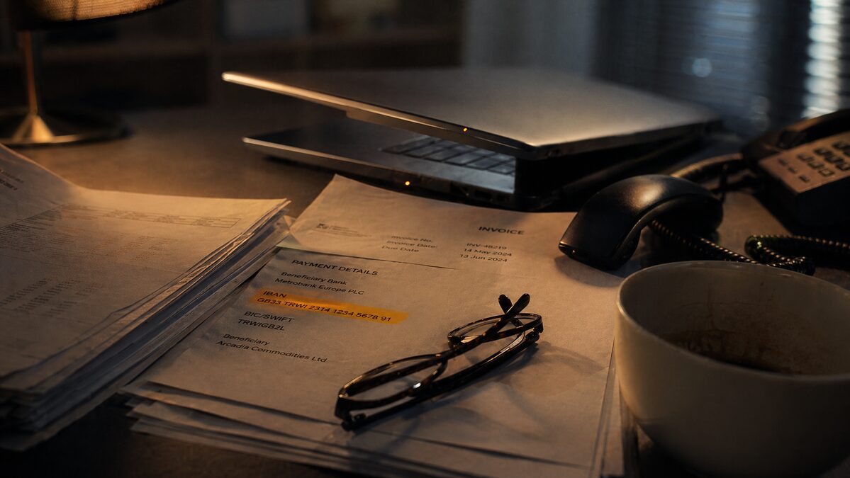 A dim office desk lit by warm tungsten light, invoices fanned out with one pulled aside revealing a substituted bank IBAN highlighted in amber, a closed laptop and an off-hook desk phone beside it.